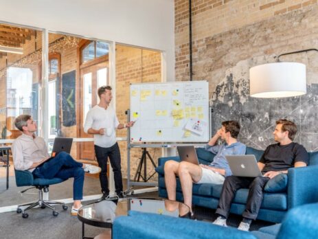 Three men using their laptops in a meeting while watching a man use a whiteboard.