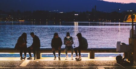 Several Gen Zers sit on a bench by the water at sundown using electronic devices. 