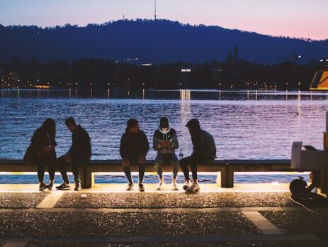 Several Gen Zers sit on a bench by the water at sundown using electronic devices. 