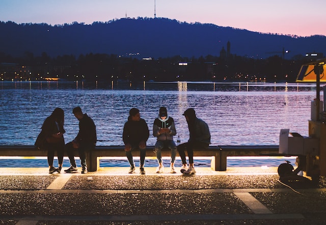 Several Gen Zers sit on a bench by the water at sundown using electronic devices. 