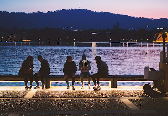 Several Gen Zers sit on a bench by the water at sundown using electronic devices. 