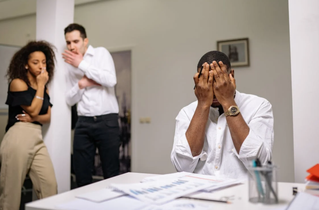 Man covering his face while two colleagues gossip behind him.