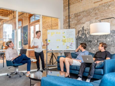 Three men sitting in an office watching a man using a whiteboard