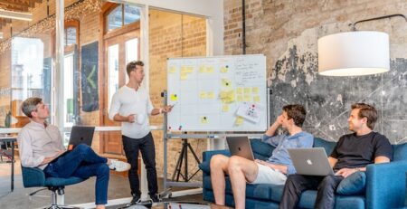 Three men sitting with laptops on their legs, and another man showing them the team goals on the board.