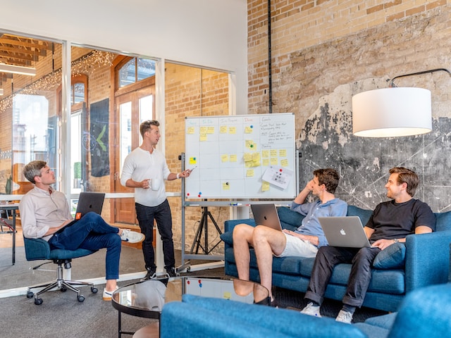 Three men sitting with laptops on their legs, and another man showing them the team goals on the board.