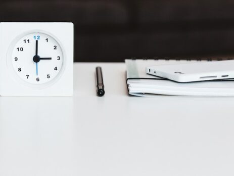 A clock placed on a table next to a pen and book