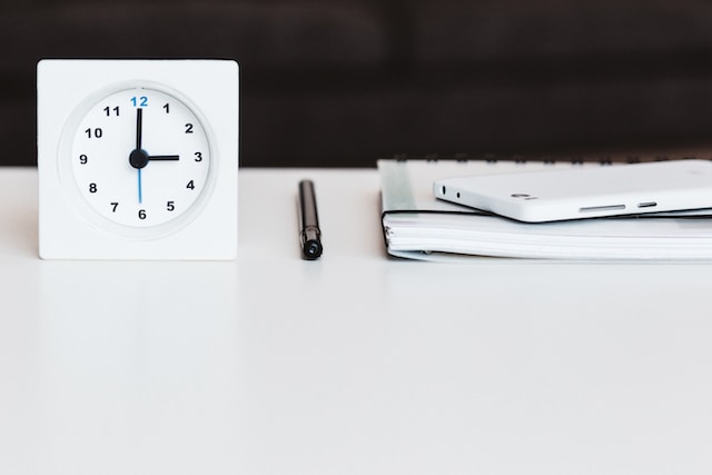 A clock placed on a table next to a pen and book