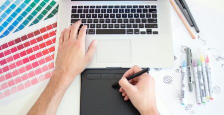 Graphic designer works with a trackpad and Macbook with color charts and markers by the sides.