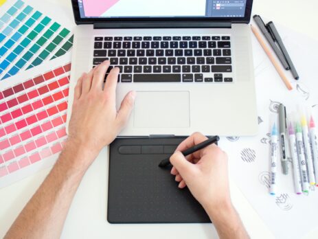 Graphic designer works with a trackpad and Macbook with color charts and markers by the sides.