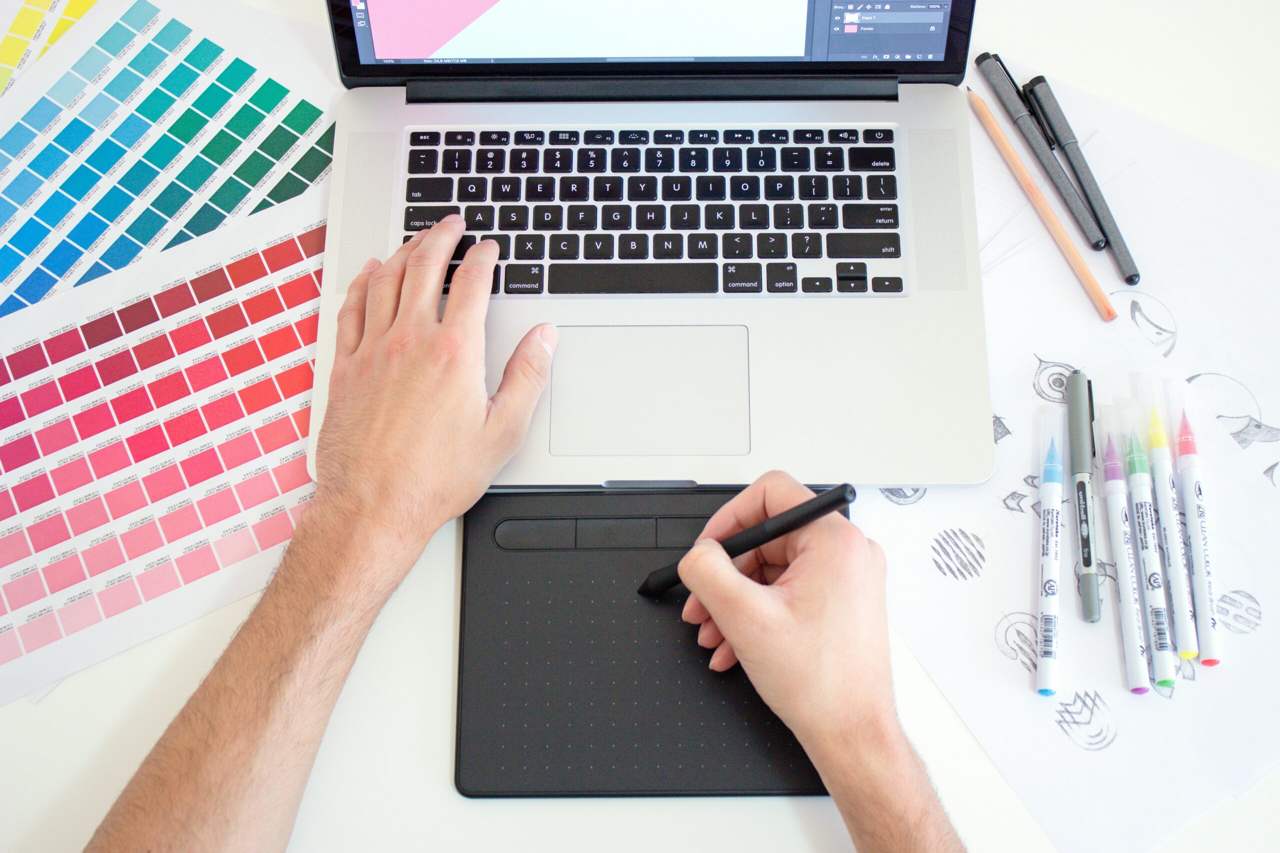 Graphic designer works with a trackpad and Macbook with color charts and markers by the sides.