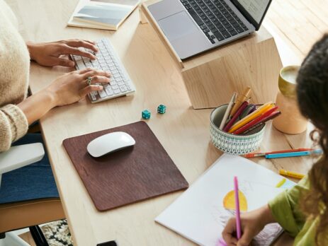 A lady works on her laptop at home while her kid makes a painting.