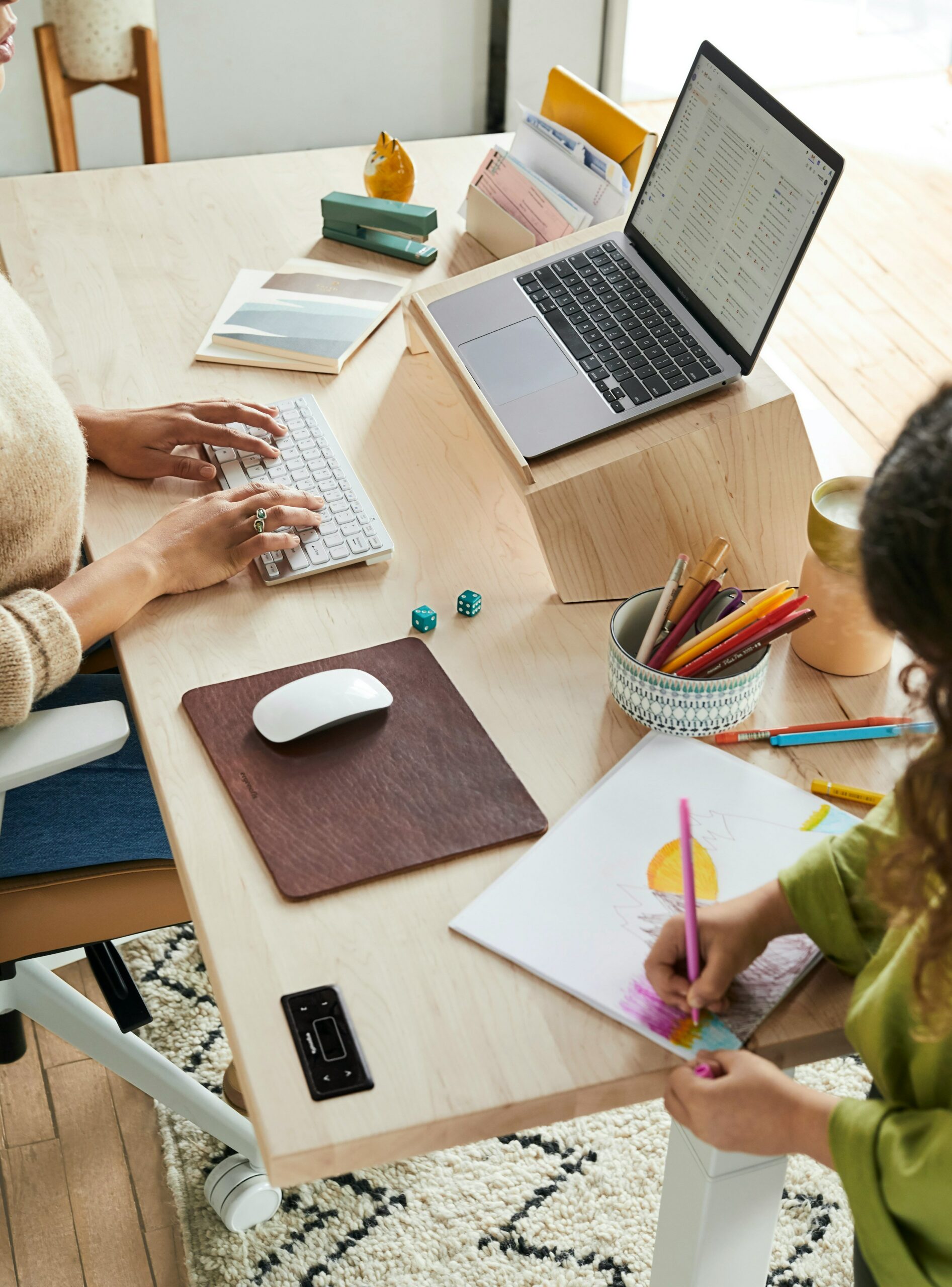 A lady works on her laptop at home while her kid makes a painting.