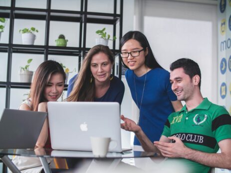 Four people watching on a white Macbook atop a glass table.