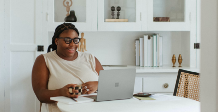 A woman smiles at her laptop while working at her kitchen table.