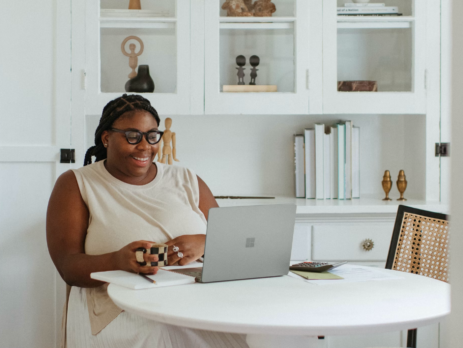 A woman smiles at her laptop while working at her kitchen table.