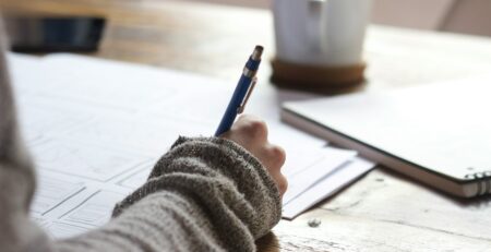 Person Writing On Brown Wooden Table Near Mug