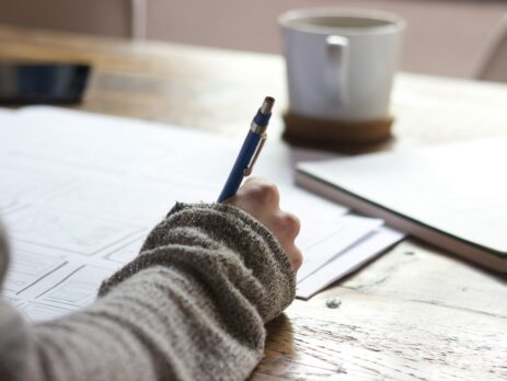 Person Writing On Brown Wooden Table Near Mug