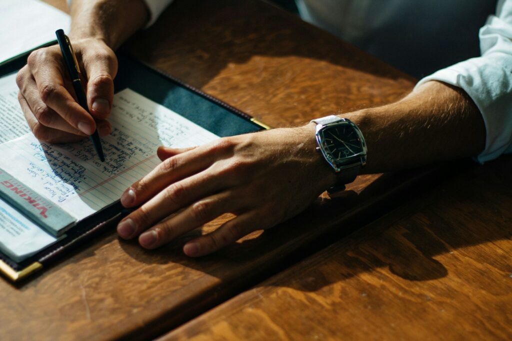 person writing in a paper on a brown table.